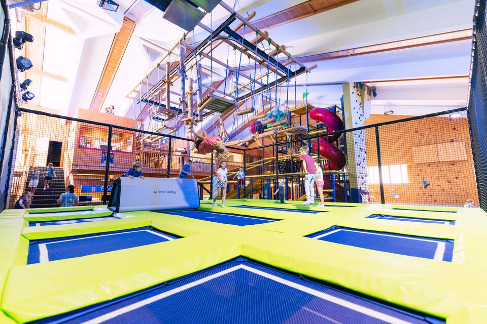 Kind schwingt an Seil in Indoor-Spielplatz mit Trampolinen und Klettergerüsten