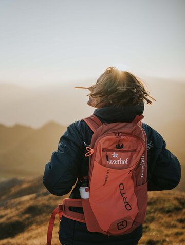 Wanderer mit rotem Rucksack blickt in die Ferne, Sonne scheint durch die Haare