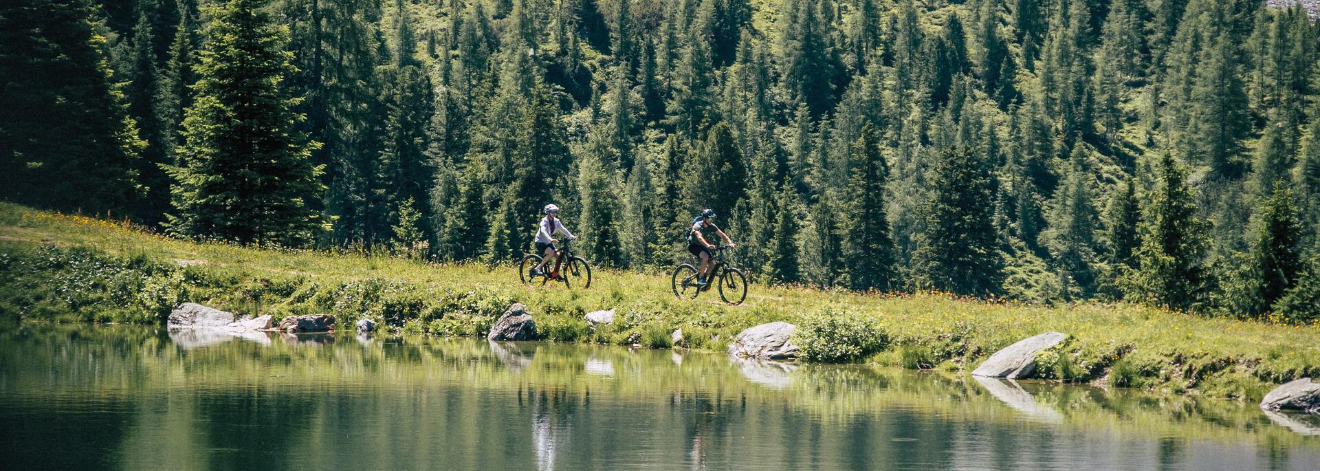 Zwei Jugendliche fahren Mountainbike am Seeufer vor bewaldetem Berghang