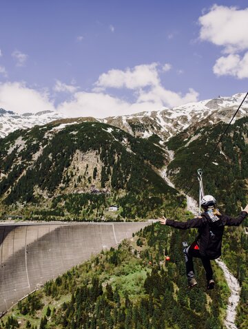 Frau fährt mit Flying Fox über Staumauer vor Bergpanorama