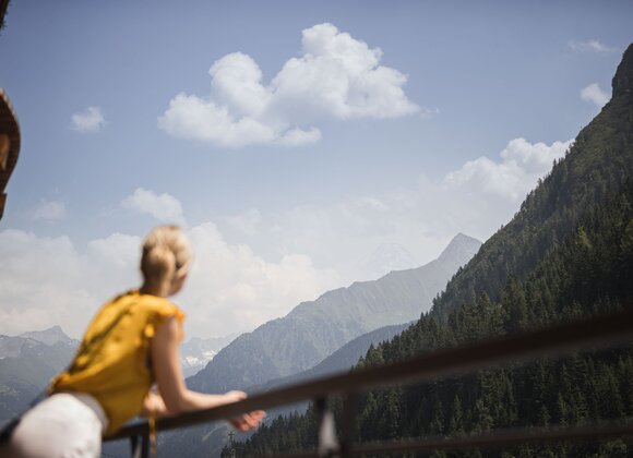 Frau blickt von Balkon auf Berglandschaft unter blauem Himmel mit Wolken
