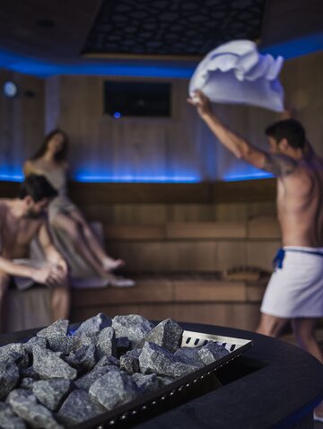 Sauna with stones in the foreground, in the background three people, one of them with a towel in the air
