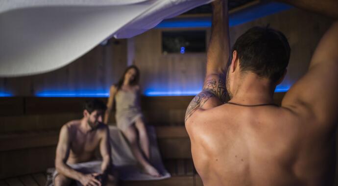 Mann hebt Tuch in Sauna, zwei weitere Personen im Hintergrund