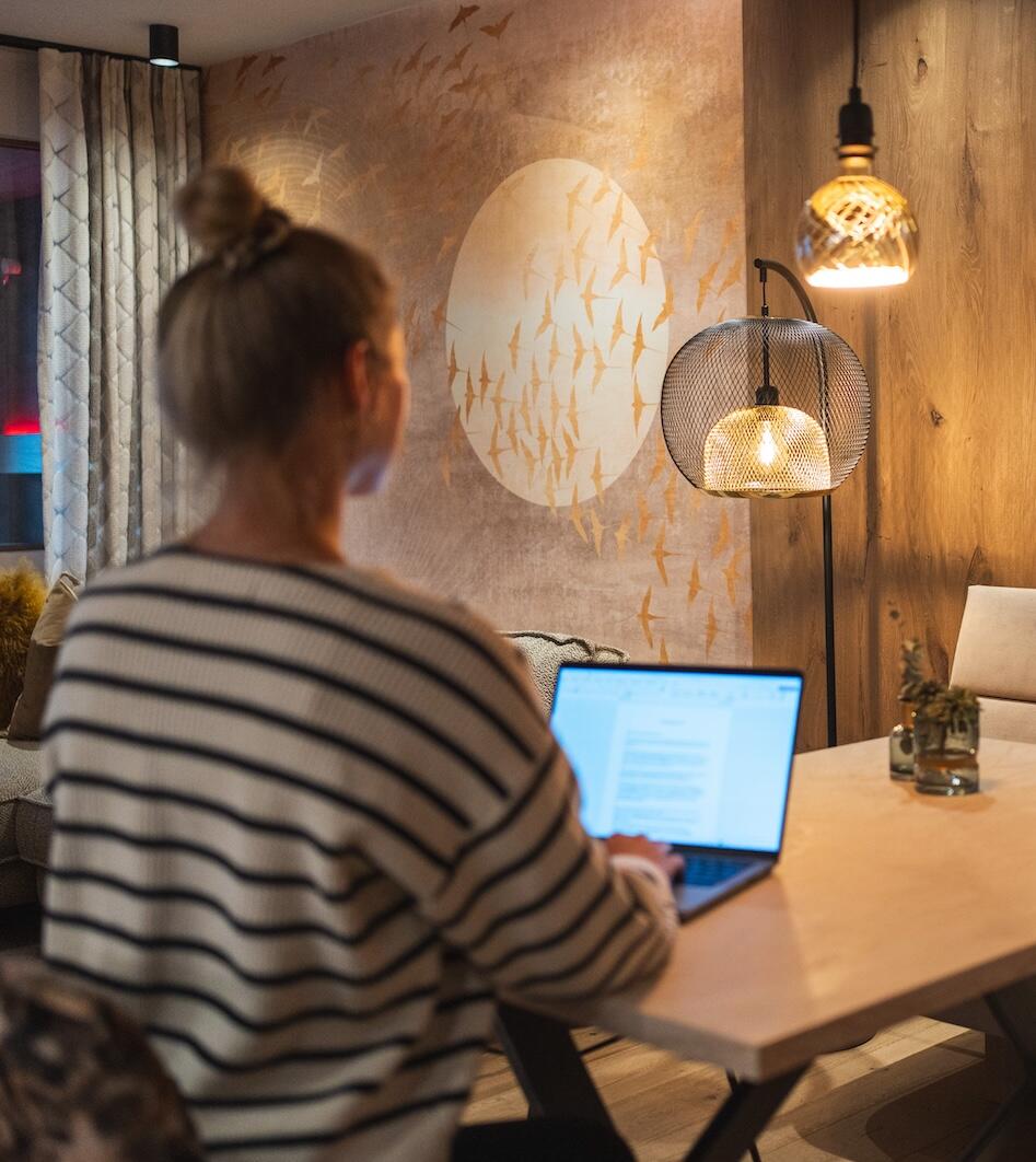 Frau mit Dutt arbeitet am Laptop in einem gemütlichen Apartment mit Stehlampe