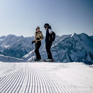 Zwei Skifahrer genießen eine präparierte Piste vor beeindruckendem Alpenpanorama
